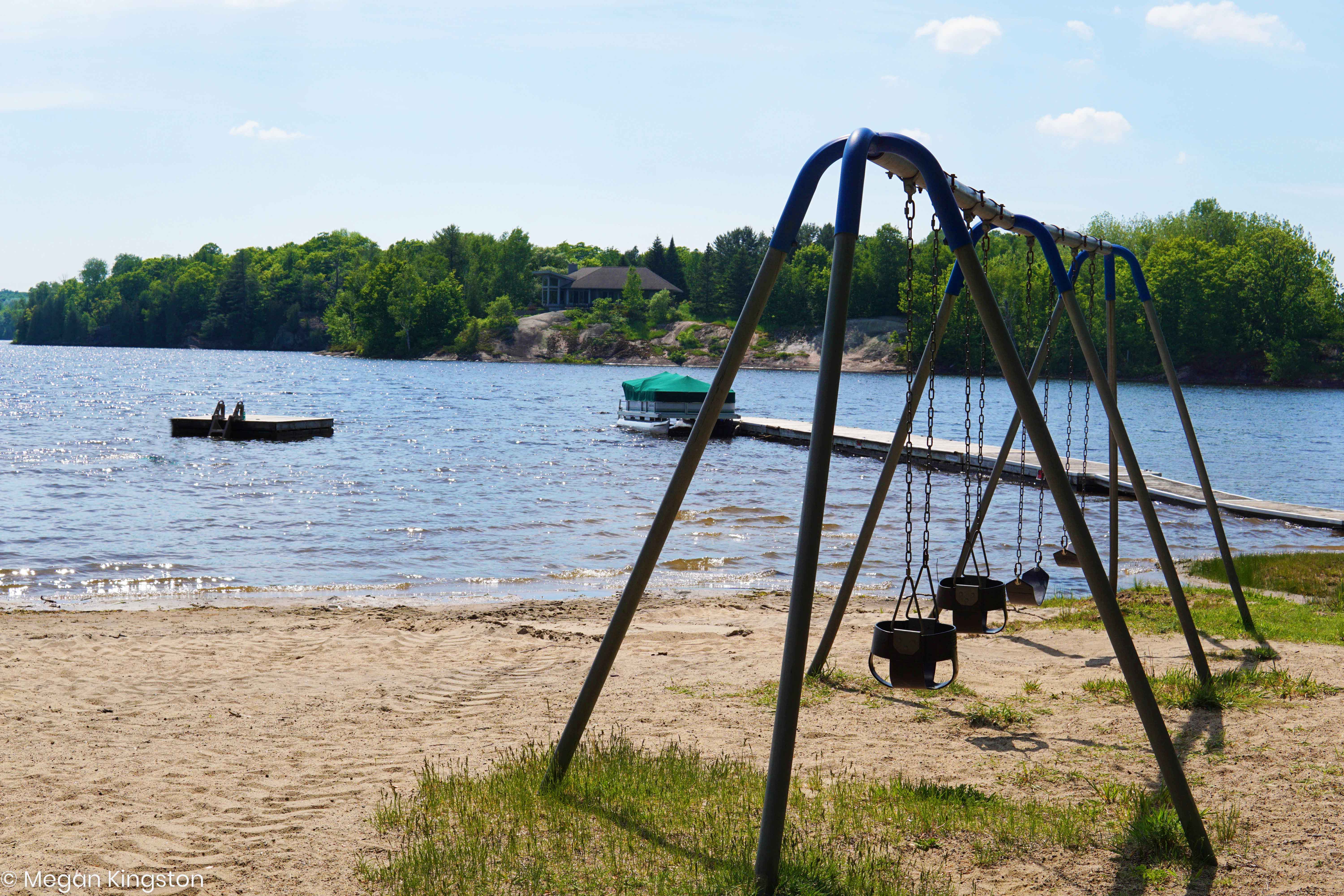 Set of swings on a sandy beach next to a beautiful lake with trees shown across the lake. A floating swim raft sits off in the distance.