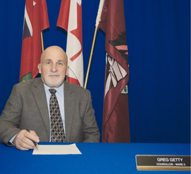 Seated man holding pen in front of Provincial, Federal and Seguin flag