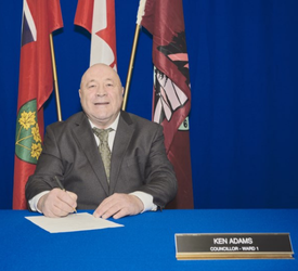 Seated man holding pen in front of Provincial, Federal and Seguin flag