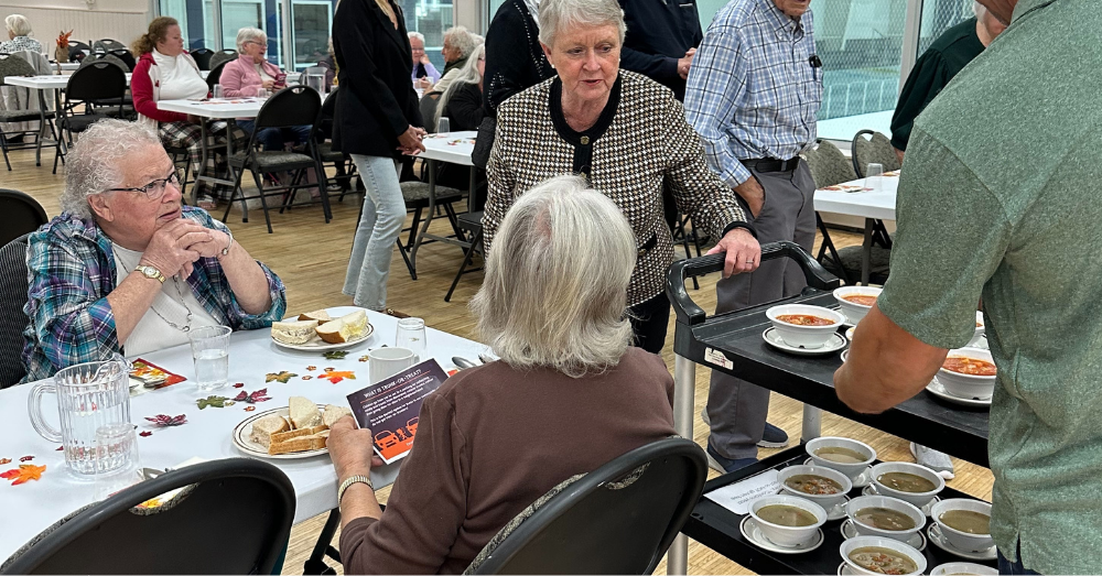 Seniors at a table with a woman serving soup from a tray