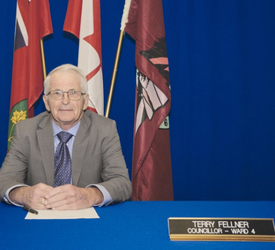 Seated man holding pen in front of Provincial, Federal and Seguin flag