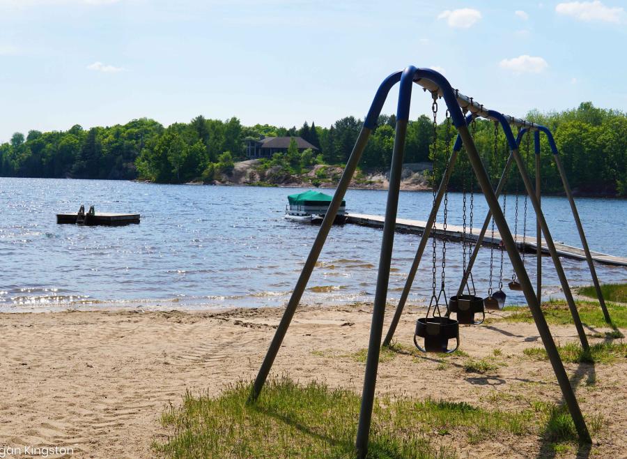 Set of swings on a sandy beach next to a beautiful lake with trees shown across the lake. A floating swim raft sits off in the distance.