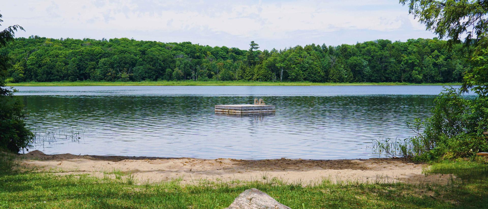 Grassy area overlooking a beautiful lake surrounded by trees on a clear summer day. A floating swim raft is seen in the centre of the lake.