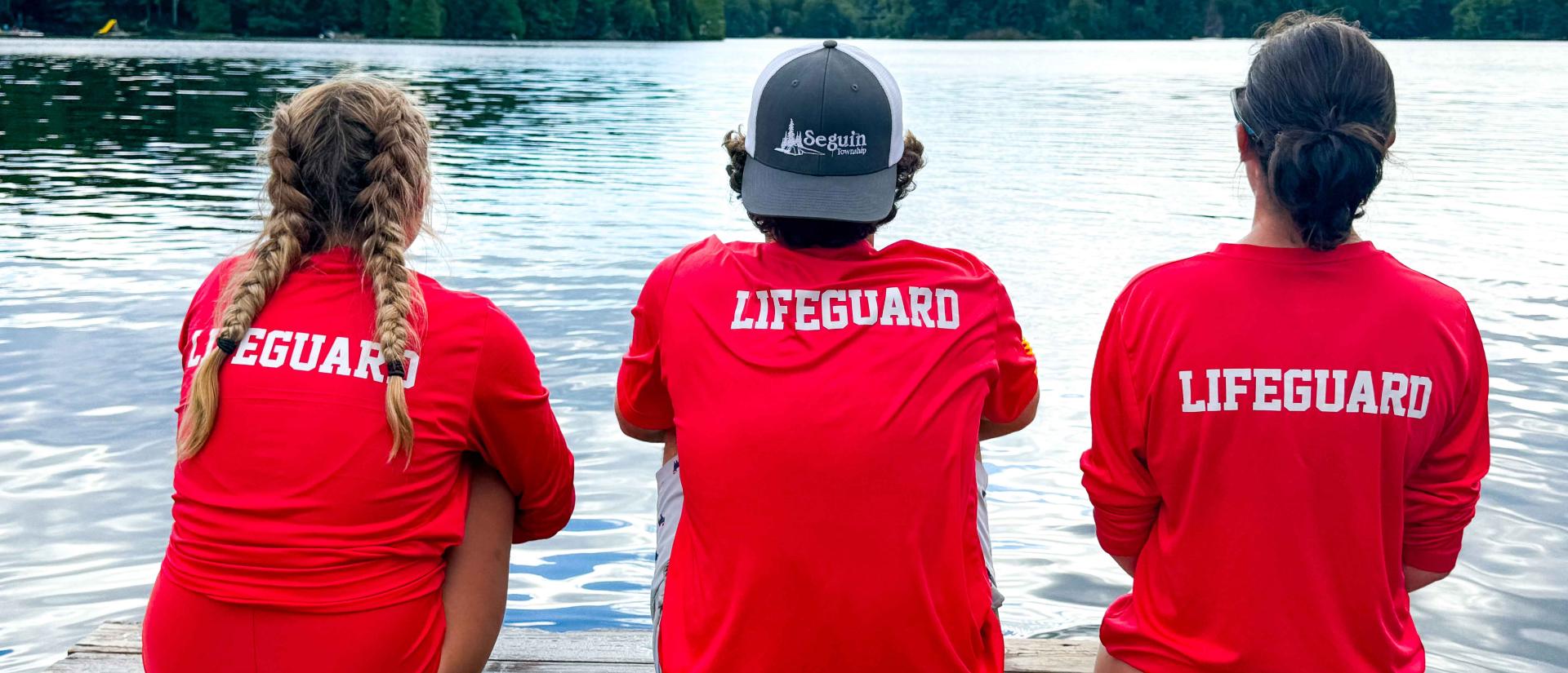 Backs of three lifeguards wearing red shirts with the word "Lifeguard" on the back, sitting on a dock overlooking a lake. The middle lifeguard is wearing a backwards hat that says "Seguin Township" on it.
