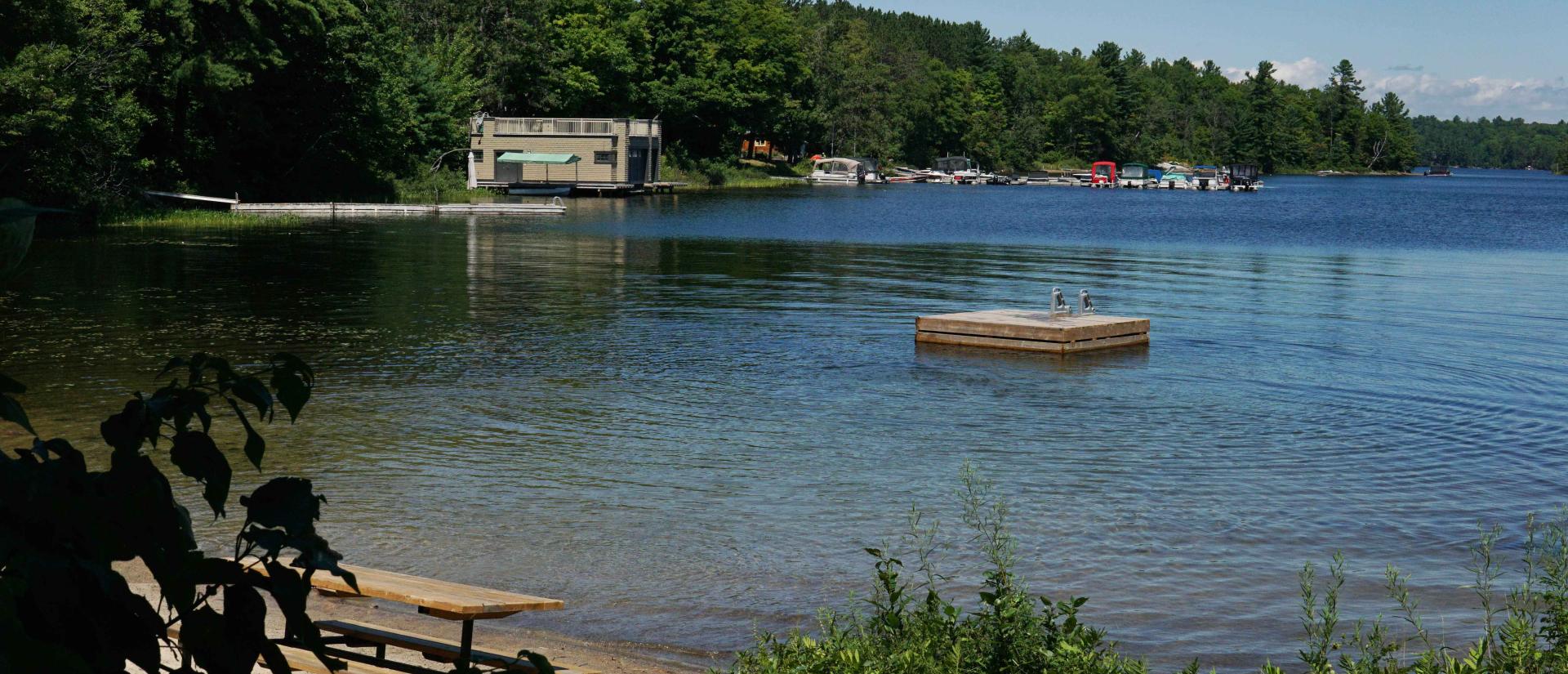 A sandy beach with a picnic table next to a clear blue lake that has a floating swim raft in the middle of it. Nearby docks and cottages are seen along the shoreline in the background.