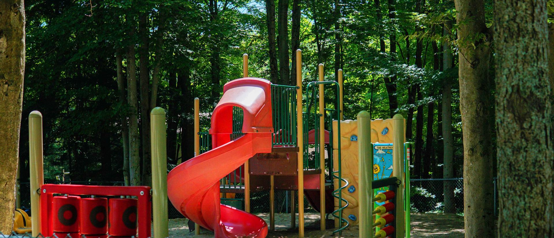 Fenced-in playground in a beautiful wooded area that features a big red slide, a giant tic-tac-toe game, and bright yellow climbing equipment.