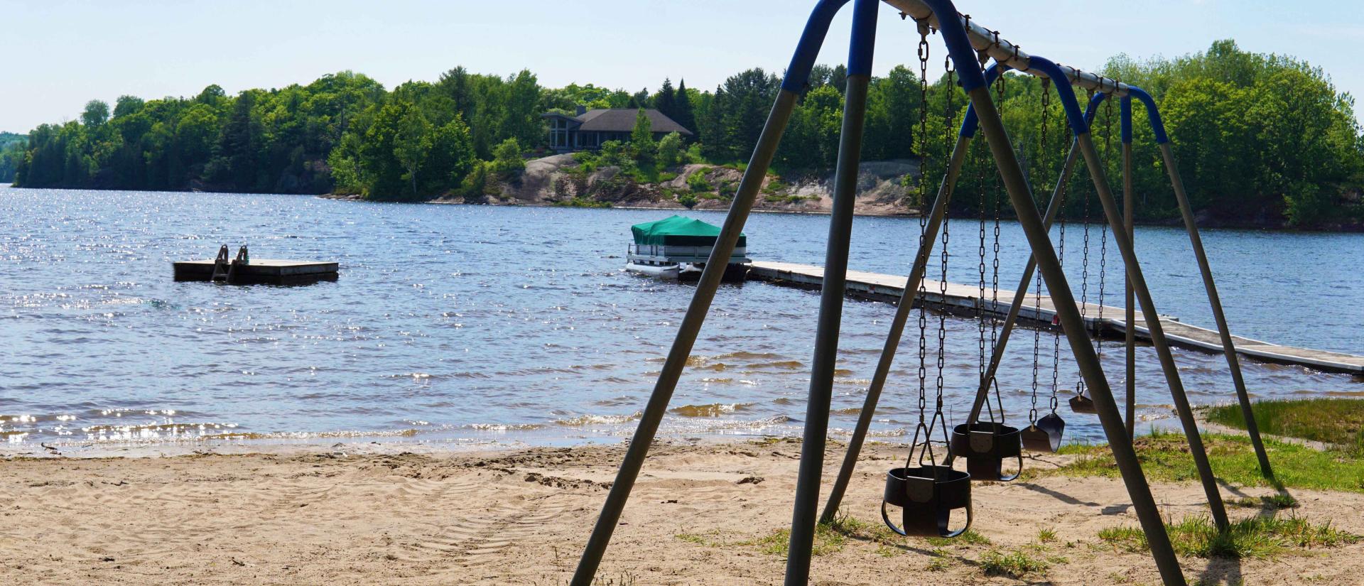 Set of swings on a sandy beach next to a beautiful lake with trees shown across the lake. A floating swim raft sits off in the distance.