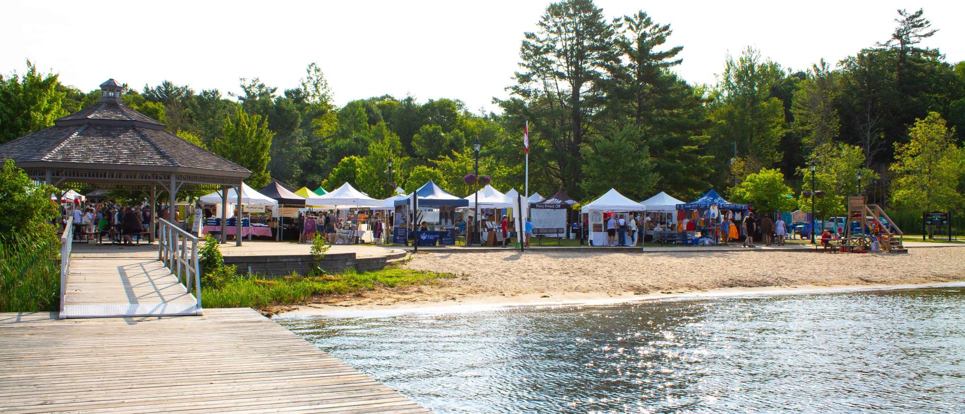 View of a beach from the dock next to a row of buoy lines in the water. A roofed pavilion and various festival tents can be seen in the distance.