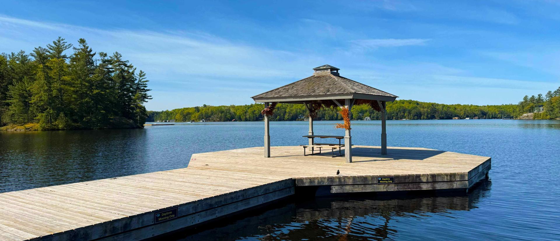 Long dock extending into the centre of a still lake on a clear summer day with trees and cottages seen in the background. A picnic table under a pavilion is at the end of the dock. 