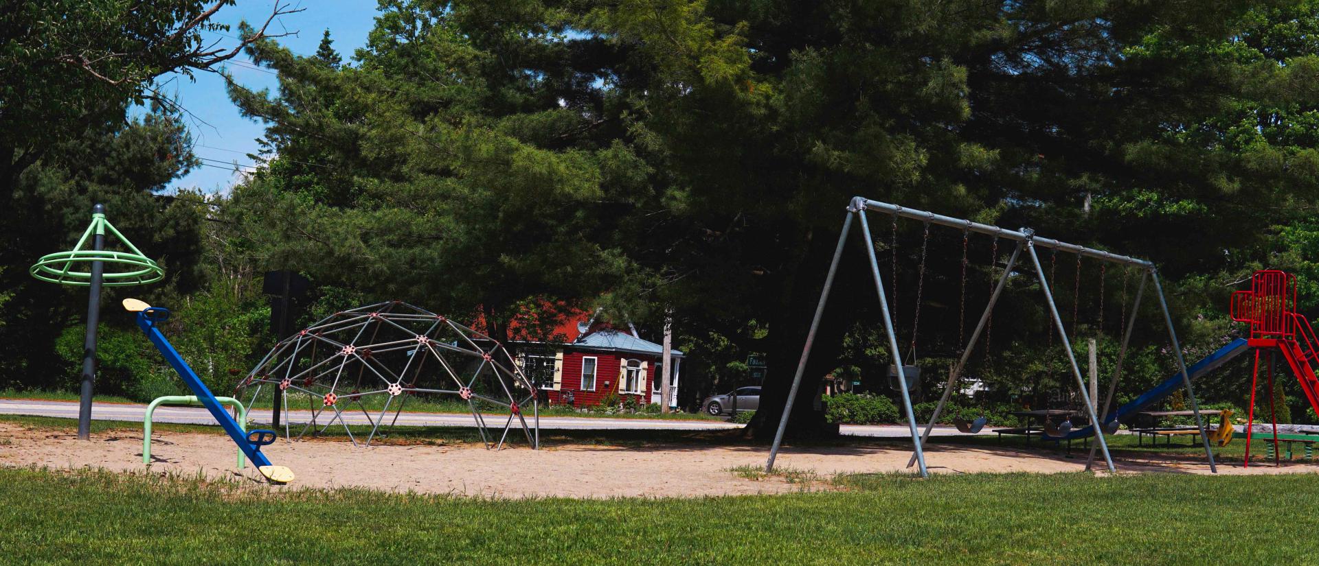 An open field with a sandy lot and playground equipment, featuring a big swing set, a teeter totter, and a carousel.