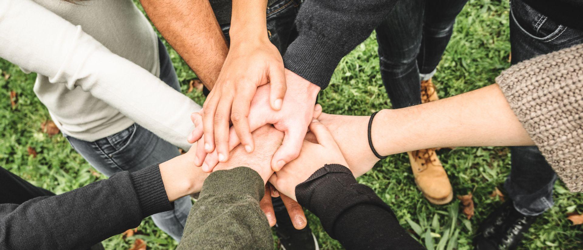 Close-up of a group of people standing in a circle outdoors on grass, stacking their hands together in the center as a symbol of teamwork and unity