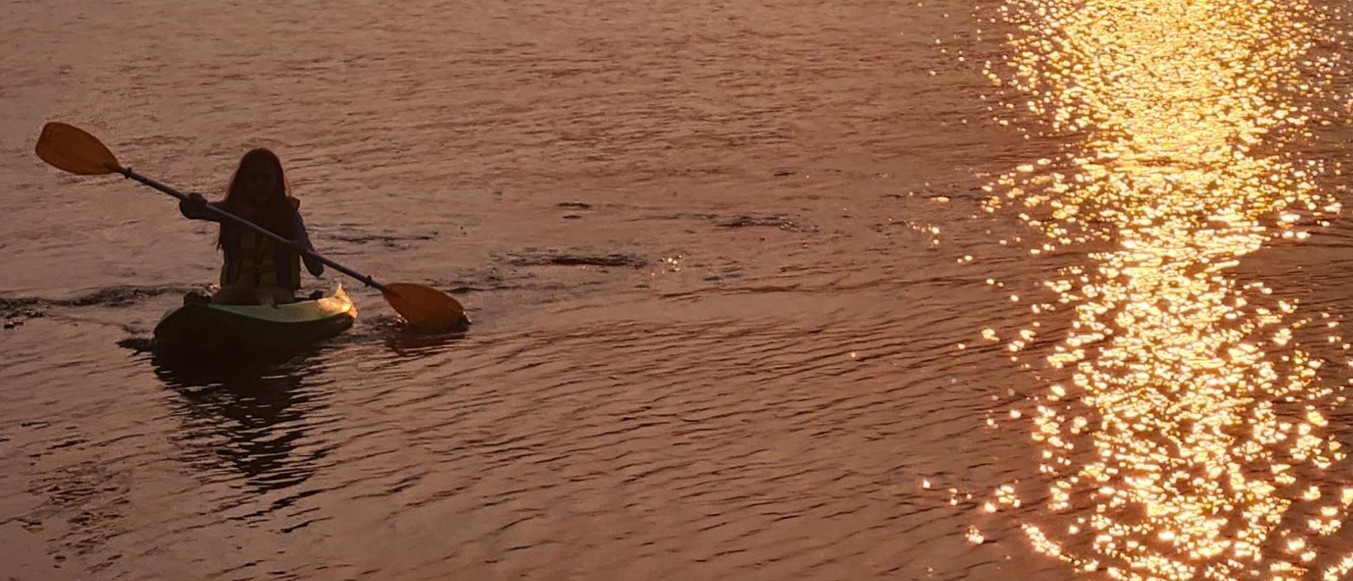 A silhouette of a woman kayaking on a lake with the sunset reflecting in the water next to her