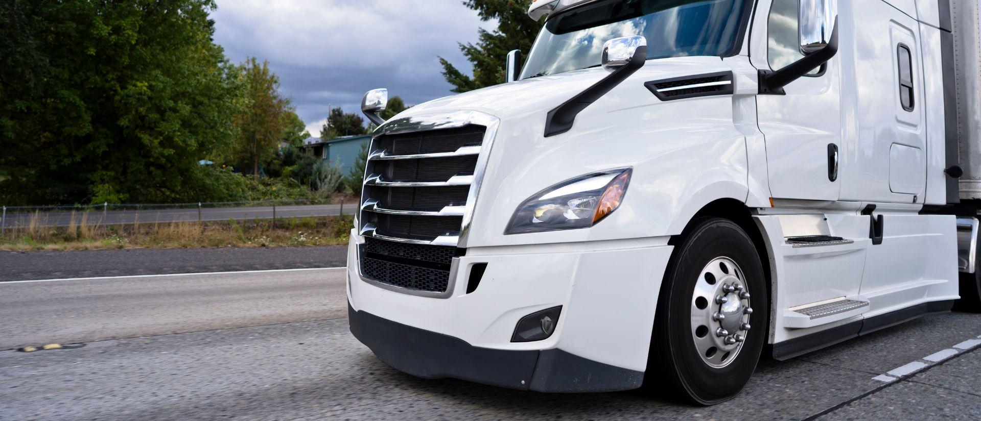 Close-up view of a white semi-truck driving on a highway with trees and cloudy sky in the background.