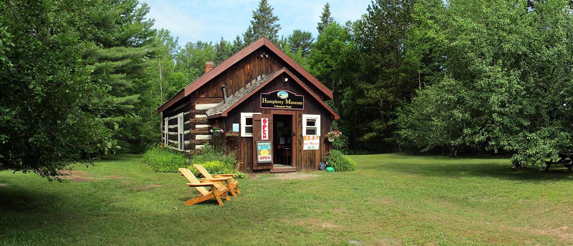 The historical Humphrey log school house museum in the summertime with an open sign and muskoka chairs sitting outside
