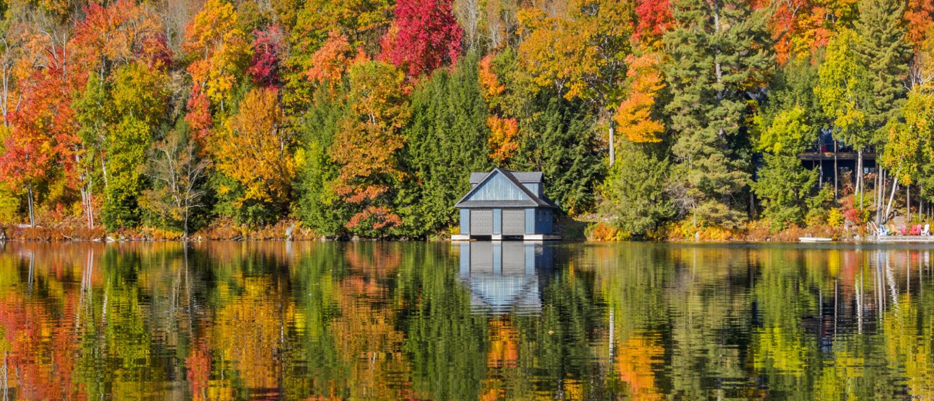 Boat house on the edge of a clear, still lake against a heavily forested backdrop of autumn trees