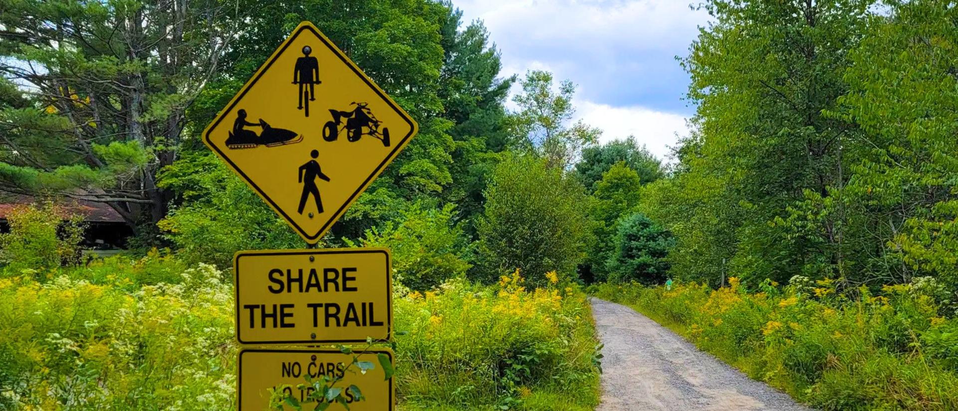 A trail in a heavily wooded area with a yellow sign next to the trail saying "Share the Trail" and showing various recreational symbols 