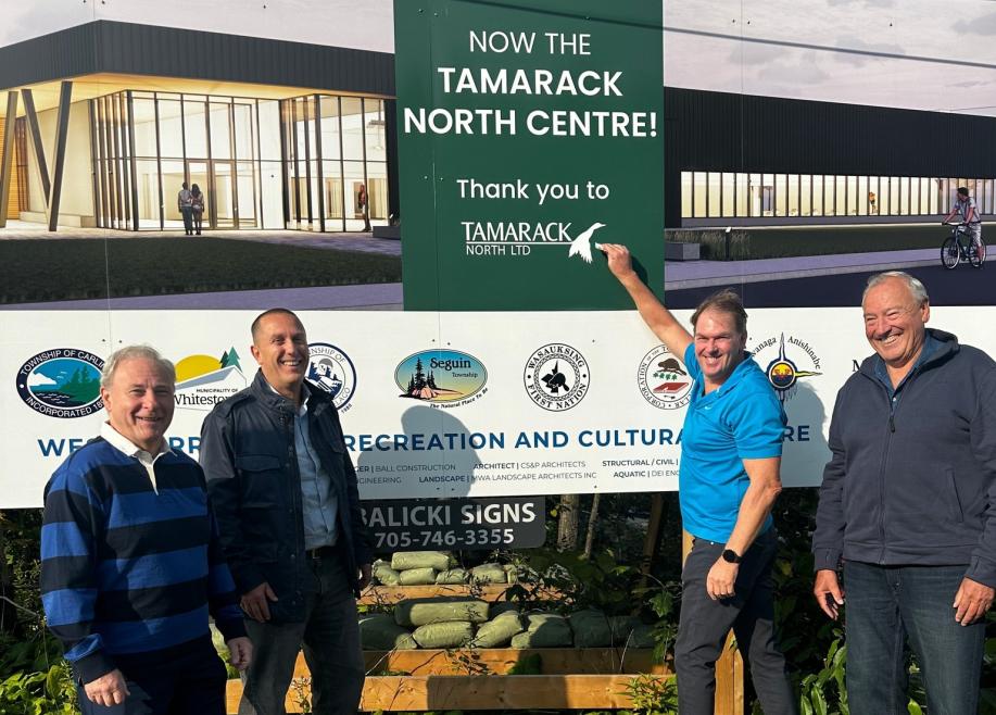Four smiling men stand in front of a sign for the new Tamarack North Centre. One points at the sign, conveying excitement and accomplishment.